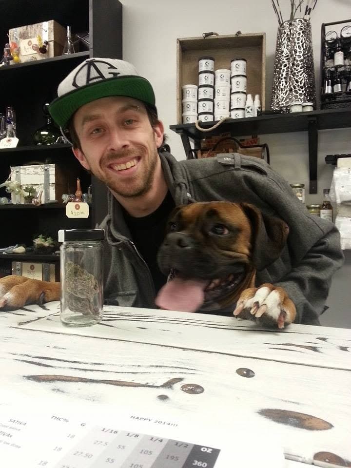 John Hirsch smiling behind a dispensary counter with a boxer dog, a jar and a price sheet in front of them.