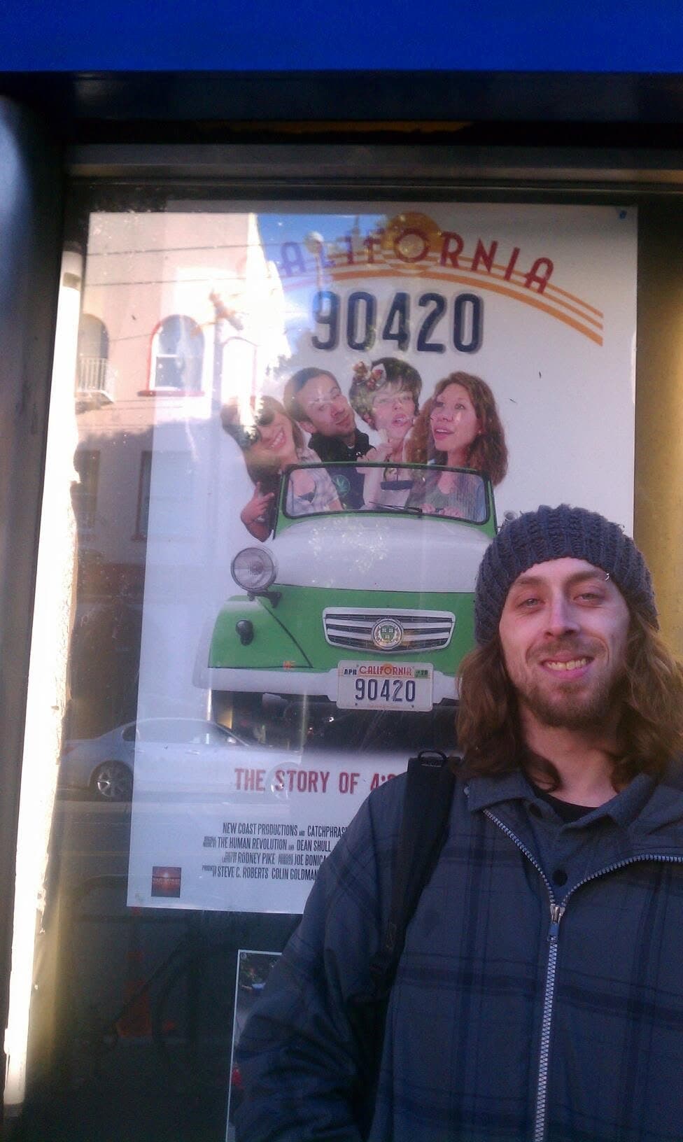John Hirsch in a beanie standing beside the California, 90420 poster in a theater display case.